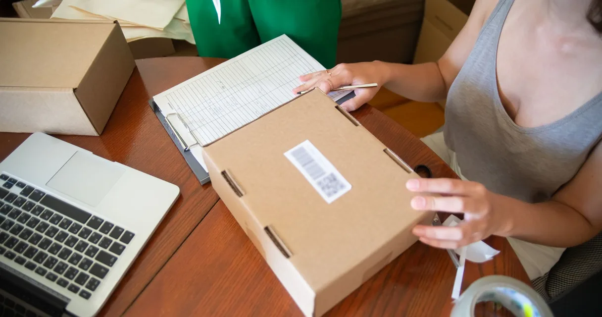 Pessoa embalando pedido em caixa de papelão com etiqueta ao lado de um notebook