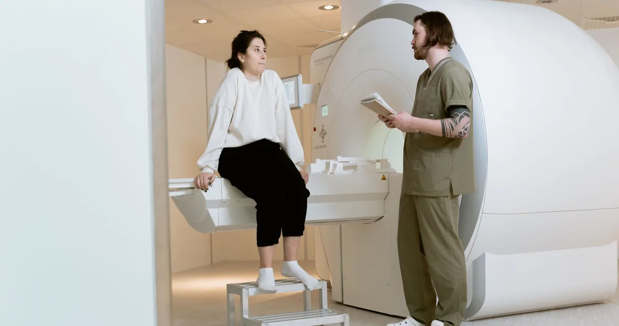 Paciente sentada na mesa do aparelho de ressonância magnética enquanto o técnico explica o exame